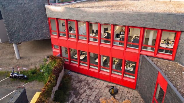 Luftaufnahme der Hofseite mit den rot gerahmten Fensterbändern über dem gepflasterten Innenhof; Büroinhalte hinter den Fenstern durch KI-Inhalte anonymisiert