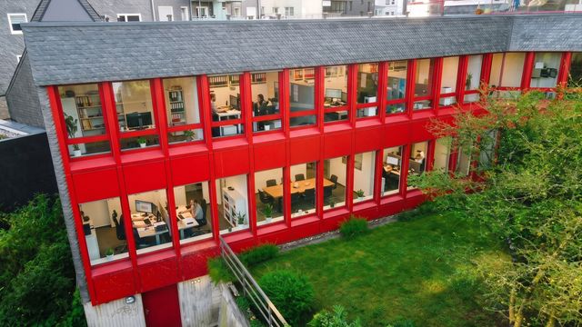 Luftaufnahme der Gartenseite mit den rot gerahmten Fensterbändern, davor Rasen und Bäume; Büroinhalte hinter den Fenstern durch KI-Inhalte anonymisiert
